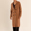A woman modeling a mid-length camel brown double-breasted coat over a dark brown V-neck top, black wide-leg trousers, and black shoes, set against a plain beige studio background.