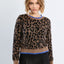 A woman modeling a cropped knit sweater with a brown and black leopard print, featuring bright blue ribbed trim at the crew neck, cuffs, and hem, paired with black trousers against a light gray studio background.