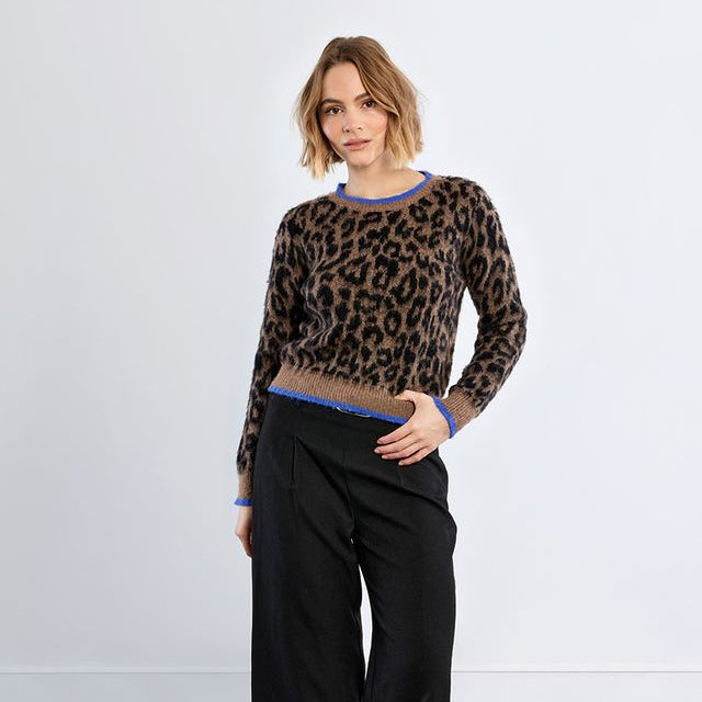 A woman modeling a long-sleeve knit sweater with a brown and black leopard print pattern and blue trim, paired with black wide-leg trousers and black leather loafers, against a light gray studio background.