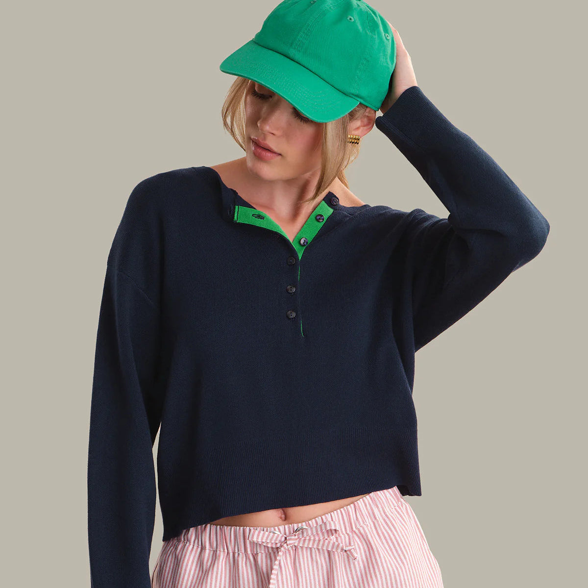 Woman wearing navy drop shoulder henley with green collar detail and green baseball cap, paired with pink striped pants Main image