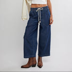 Woman wearing dark blue denim pull-on pants with white drawstring waist, styled with white crop top and brown leather cowboy…