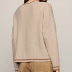 A woman shown from the back wearing a light beige knit sweater with brown striped ribbed trim at the hem and cuffs, paired with brown pants, against a plain light gray studio background.