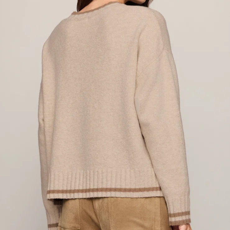 A woman shown from the back wearing a light beige knit sweater with brown striped ribbed trim at the hem and cuffs, paired with brown pants, against a plain light gray studio background.