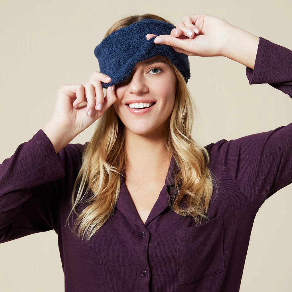 Woman wearing purple button-up shirt adjusting navy blue soft marshmallow eye mask over her eyes while smiling