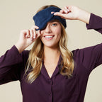 Woman wearing purple button-up shirt adjusting navy blue soft marshmallow eye mask over her eyes while smiling