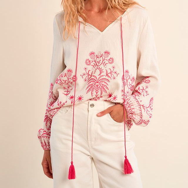 Woman wearing a white blouse with pink floral embroidery and tassels on a beige background