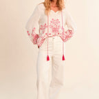 Woman wearing a white blouse with pink floral embroidery and white pants on a beige background