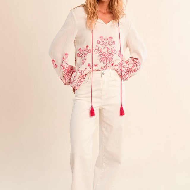 Woman wearing a white blouse with pink floral embroidery and white pants on a beige background