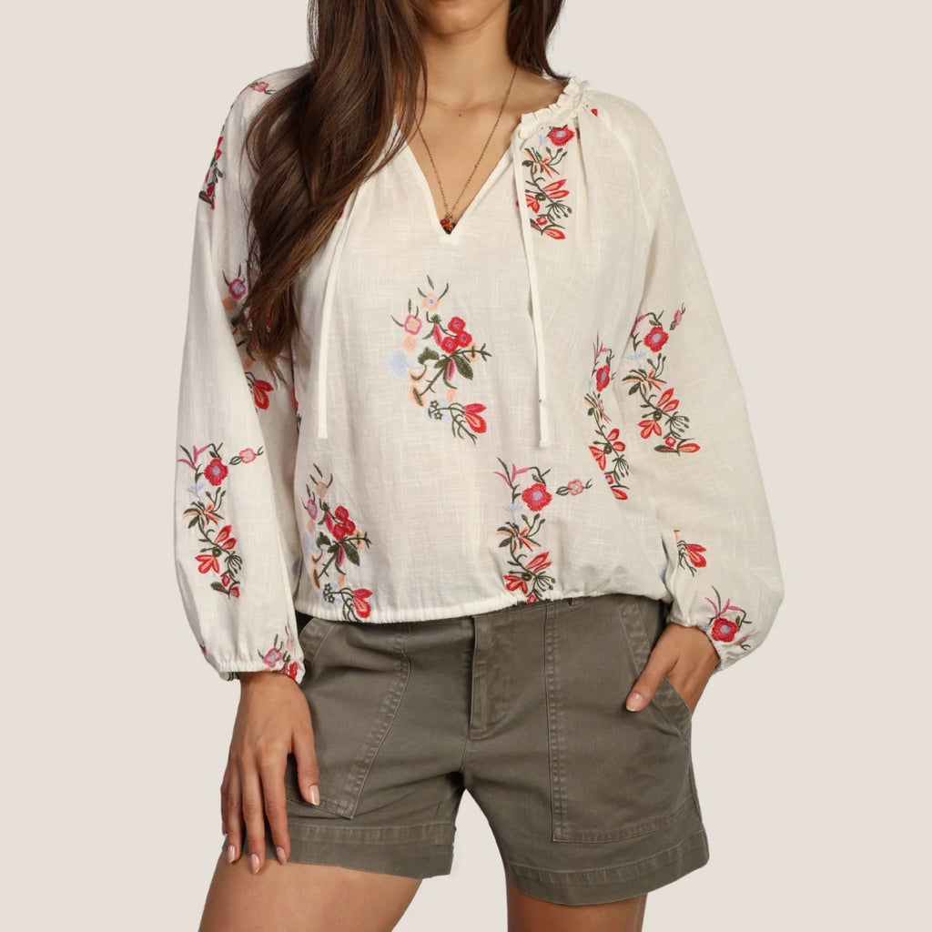 Woman wearing a white blouse with floral embroidery and green shorts on a plain background