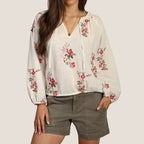 Woman wearing a white blouse with floral embroidery and green shorts on a plain background
