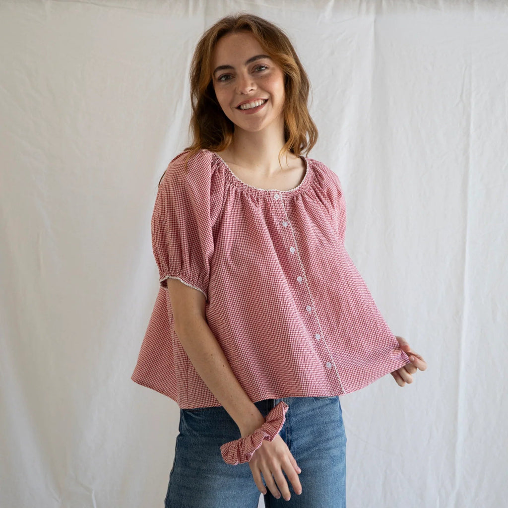 Woman wearing pink gingham Joey Blouse with short puffed sleeves and button front, styled with blue jeans