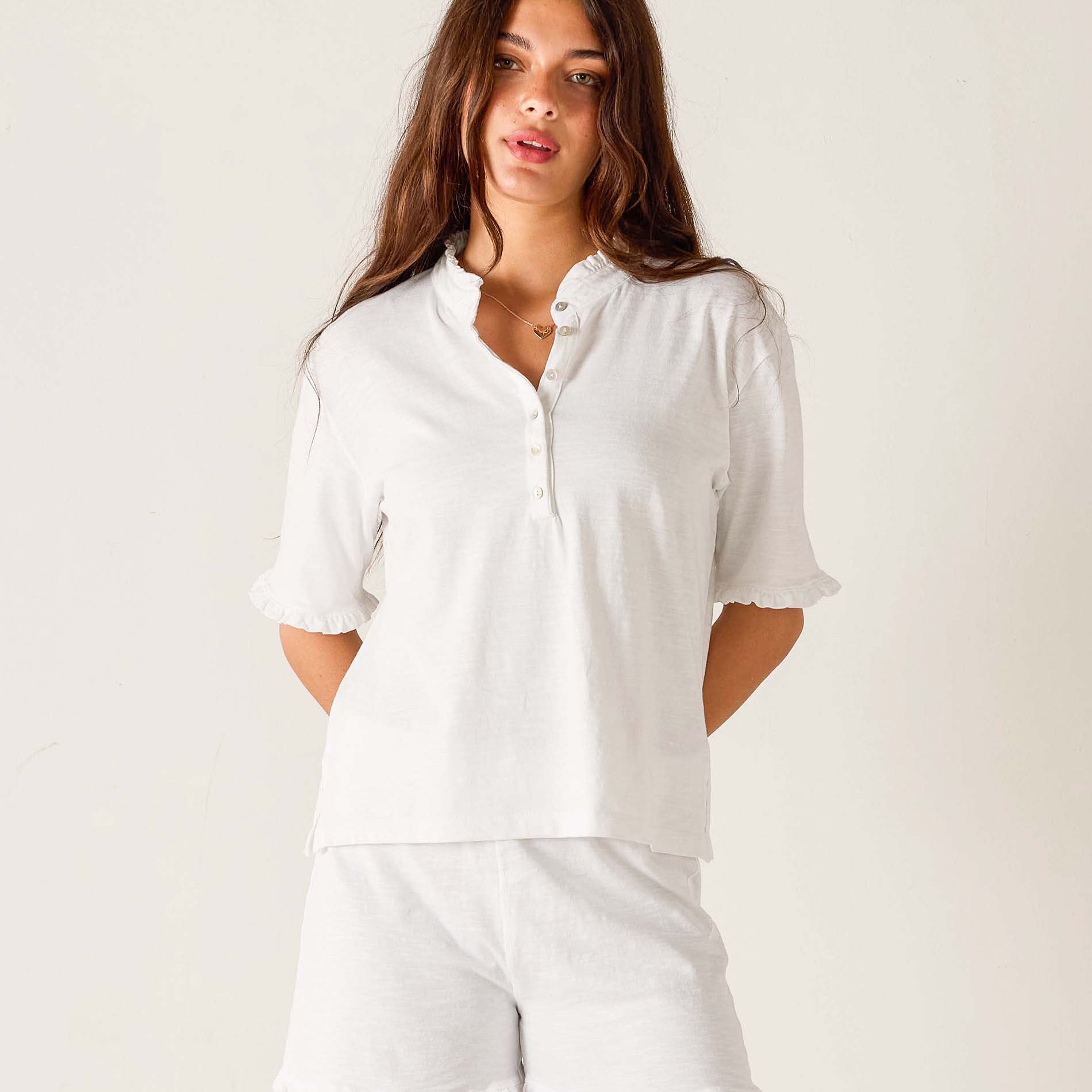 Woman wearing white Josephine Ruffle Henley Tee with half sleeves and button front, paired with white pants