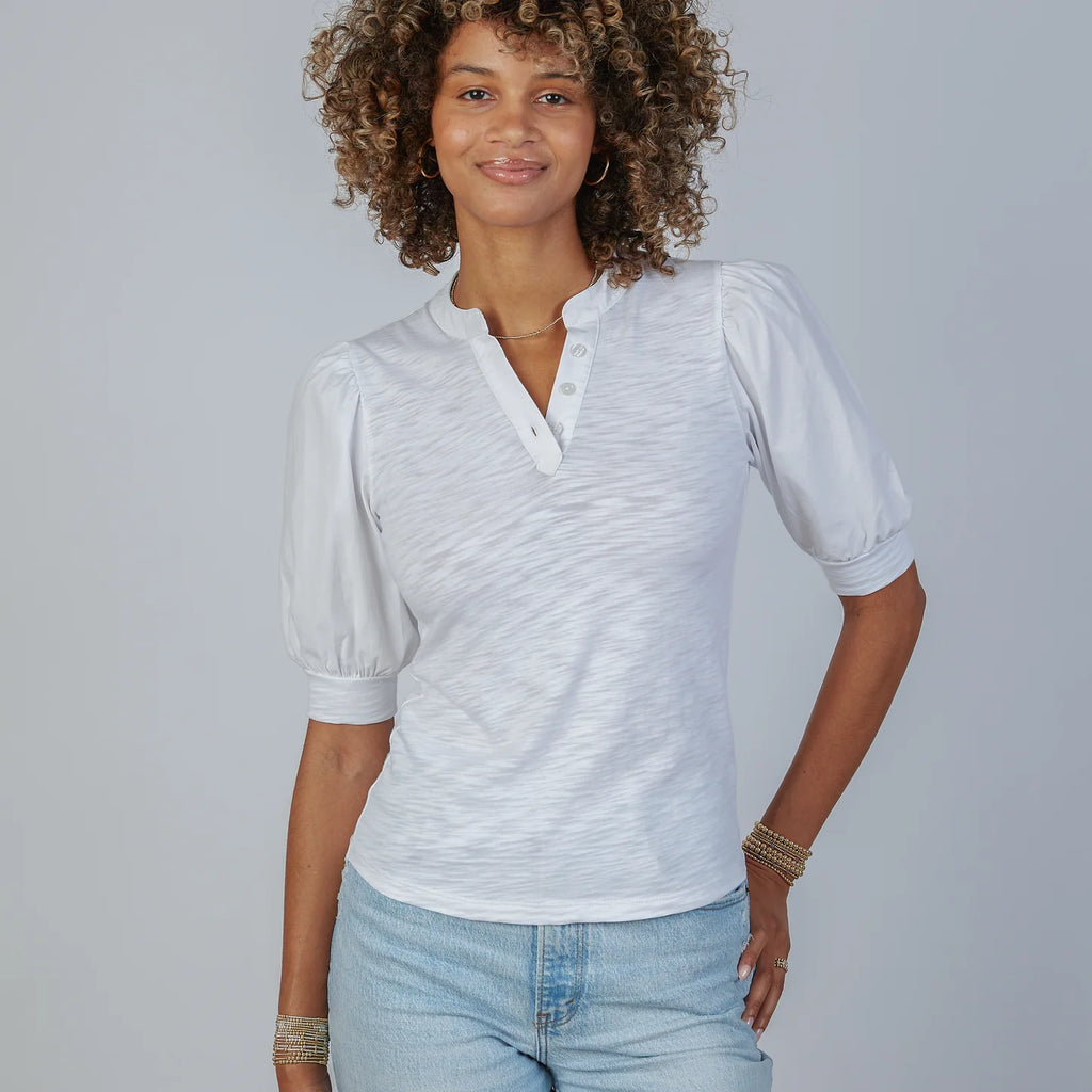 Woman wearing white Kate Puff Sleeve V-Neck Slub Tee with light blue jeans and gold bracelets, standing against gray backgro…
