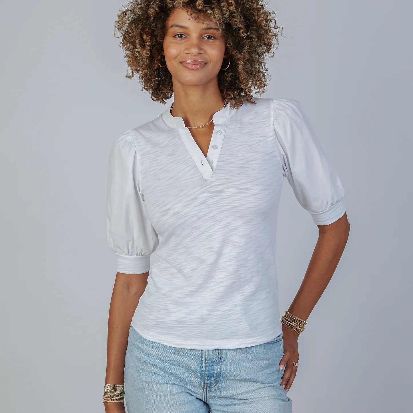Woman wearing white Kate Puff Sleeve V-Neck Slub Tee with light blue jeans and gold bracelets, standing against gray backgro…