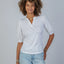 Woman wearing white Kate Puff Sleeve V-Neck Slub Tee with light blue jeans and gold bracelets, standing against gray backgro…