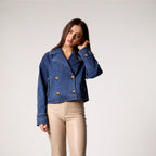 Woman wearing blue double-breasted denim cropped trench jacket with copper buttons and rolled sleeves, paired with cream-col…