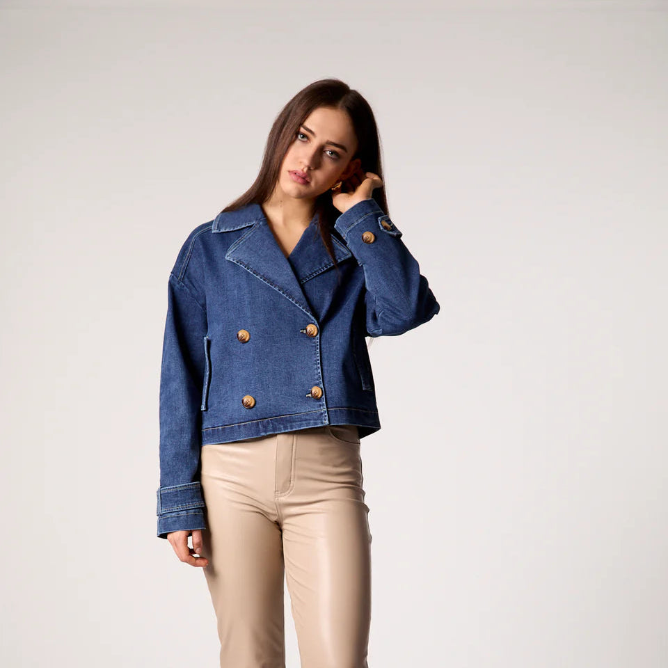 Woman wearing blue double-breasted denim cropped trench jacket with copper buttons and rolled sleeves, paired with cream-col… Main image