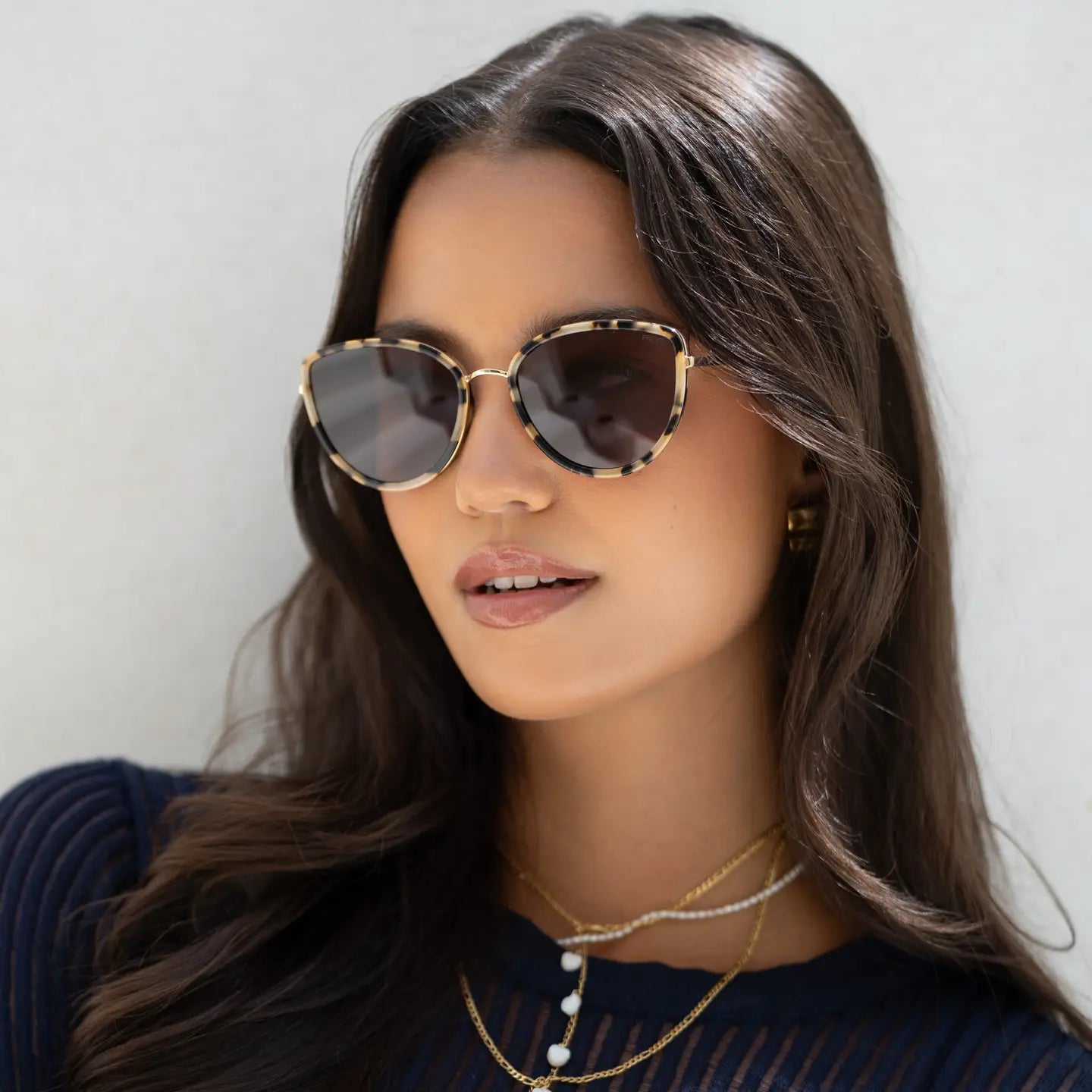 Woman wearing Luna tortoiseshell cat-eye sunglasses with gold-tone metal frames and dark lenses, styled with layered gold ne…