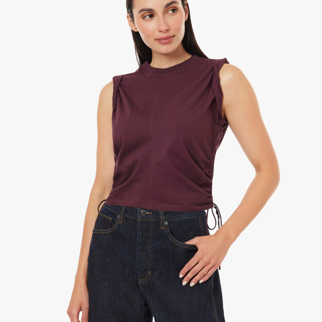 A woman modeling a sleeveless, mock-neck top in deep burgundy, paired with dark blue jeans, against a plain white studio background.