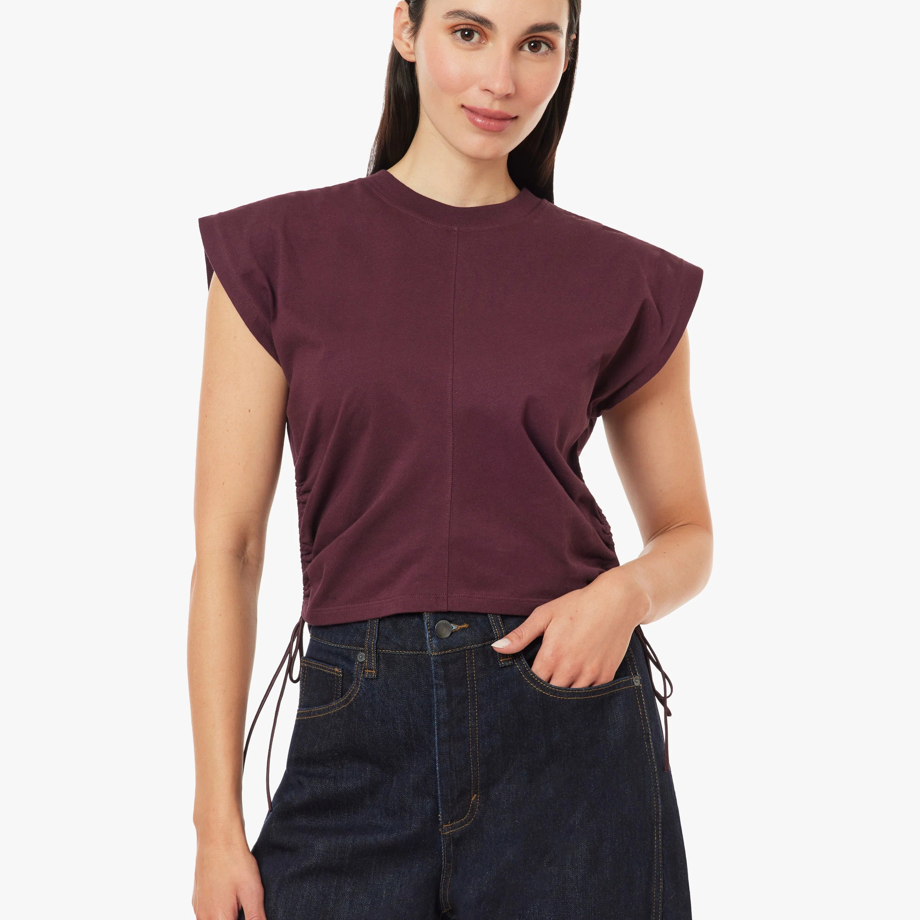 A woman modeling a dark plum purple cropped t-shirt with wide, slightly ruffled cap sleeves, paired with dark blue jeans, against a solid off-white background.