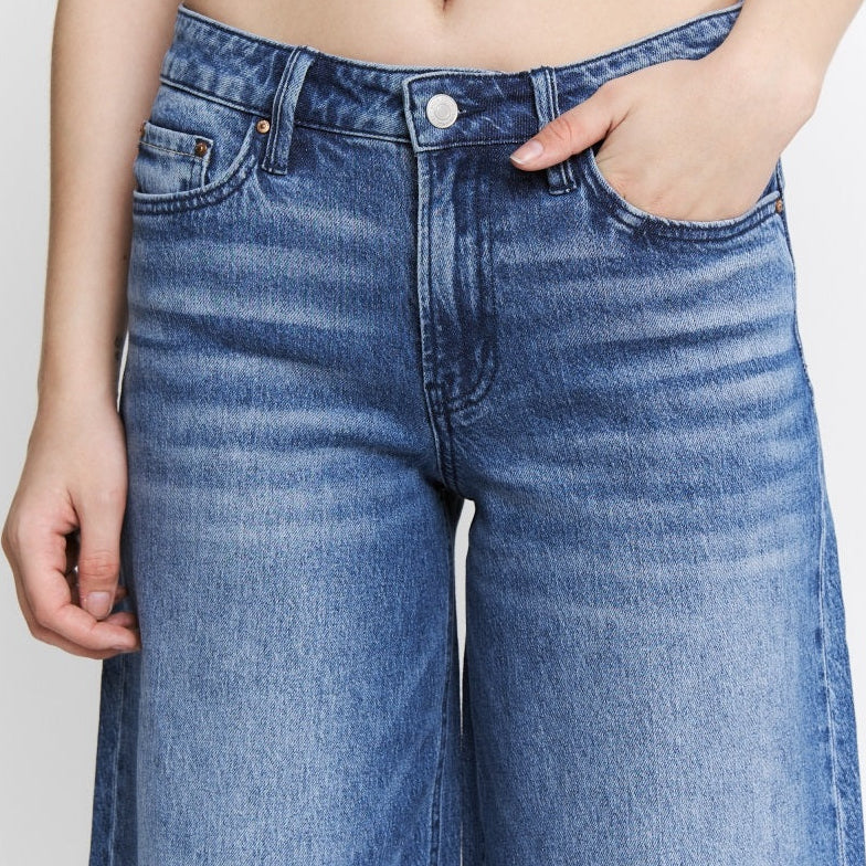 Mica High Rise Wide Leg Jeans in medium blue wash, shown on model with hands in pockets, featuring classic five-pocket styli…