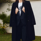 A woman stands outdoors in front of a white wall and greenery, wearing a long dark navy coat over a dark pinstripe suit with a white graphic T-shirt.