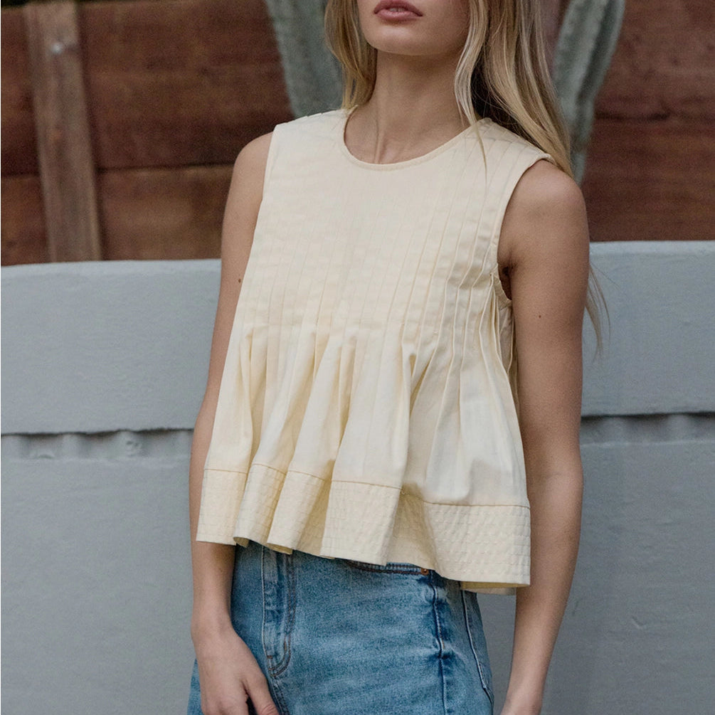 Woman wearing cream sleeveless pleated peplum top with flare hem, paired with blue denim skirt
