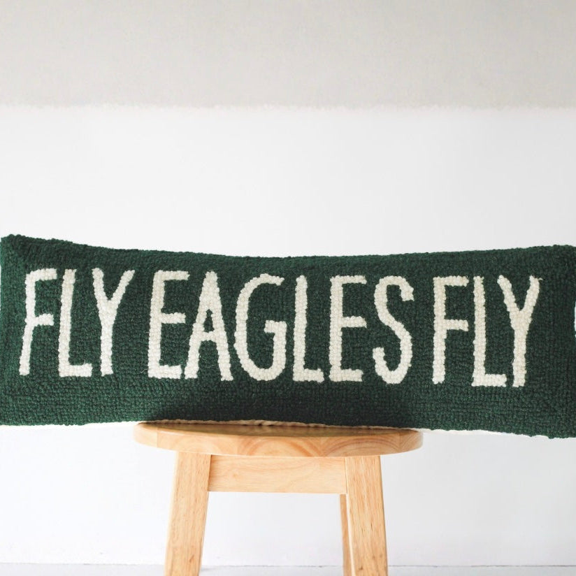 Dark green knit pillow with cream embroidered text reading Fly Eagles Fly, displayed on a wooden stool against white wall Main image