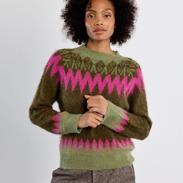 A woman with curly dark hair wearing a fuzzy olive green knit sweater with bright pink geometric patterns and brown trousers, standing against a plain light gray studio background.