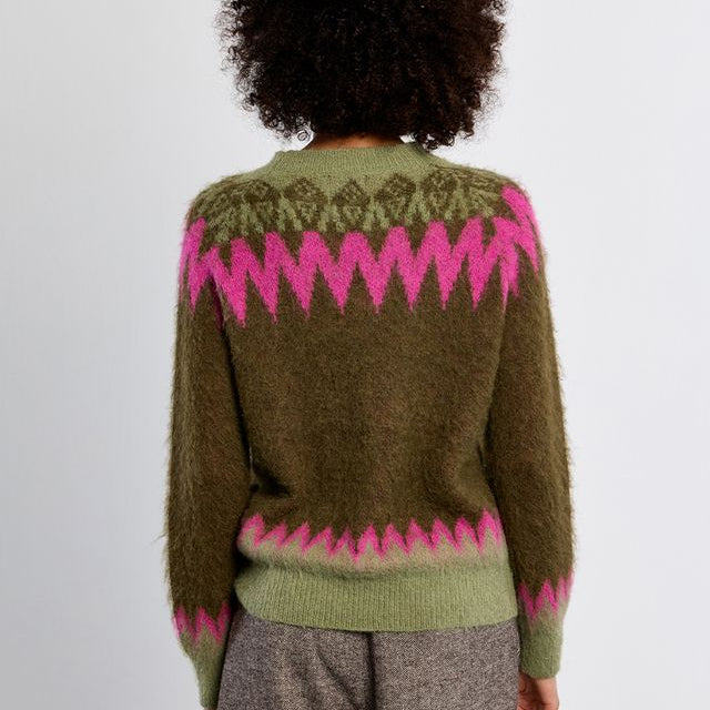 A person viewed from the back wearing a fuzzy olive green sweater with bright pink zigzag Fair Isle patterns across the shoulders, upper back, and hem, paired with taupe-gray trousers against a plain light gray wall.