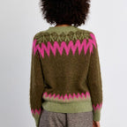 A person viewed from the back wearing a fuzzy olive green sweater with bright pink zigzag Fair Isle patterns across the shoulders, upper back, and hem, paired with taupe-gray trousers against a plain light gray wall.