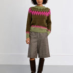 A model stands against a light gray studio background wearing a fuzzy olive green sweater with a pink and dark green Fair Isle yoke pattern, paired with gray textured knee-length shorts and dark brown knee-high boots.