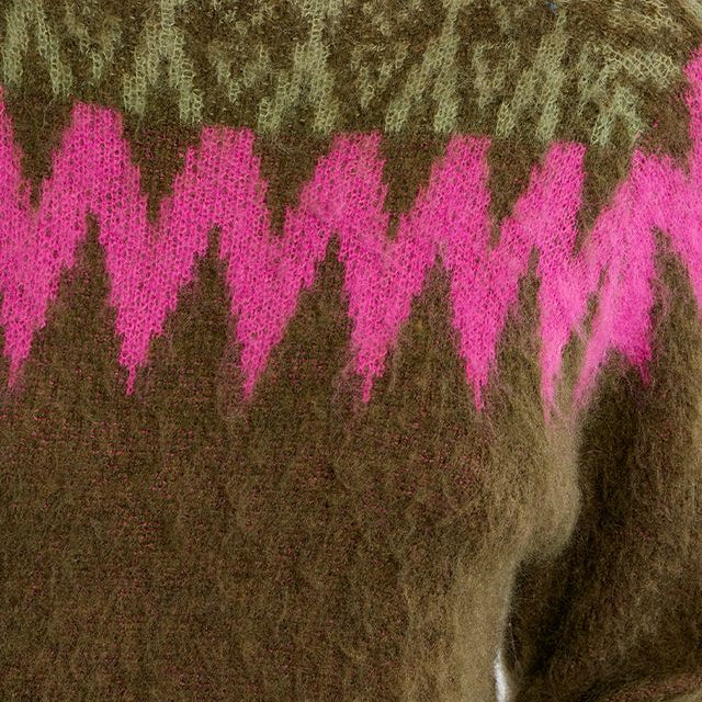 Close-up of a fuzzy knit sweater with a bright pink zigzag pattern across an olive green base, featuring a darker olive yoke with a geometric pattern.