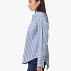 A woman shown in side profile wearing a light blue button-up shirt with thin vertical dark stripes and dark blue jeans, photographed against a plain light gray studio background.
