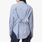 A woman shown from the back wearing a light blue and white vertically striped button-up shirt with a tie detail at the waist, paired with dark blue jeans, against a plain white background.