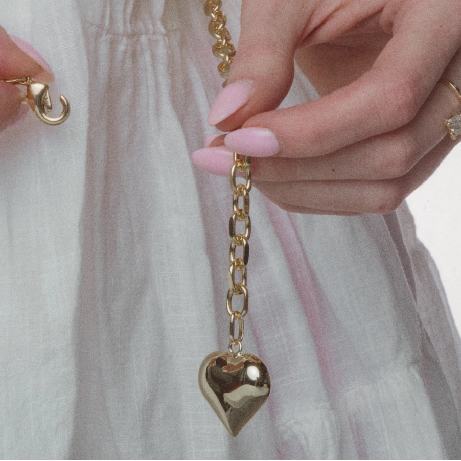 A close-up of a person wearing a white textured dress, holding a gold heart-shaped pendant necklace on a delicate gold chain. The person has light pink manicured nails and is wearing pearl and gold bracelets on one wrist and a gold ring on the other hand.
