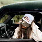 Person wearing a camo and white cap with text "Strong as a Mother" inside a car, looking out the window.
