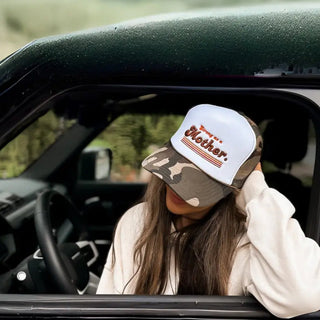 Person wearing a camo and white cap with text "Strong as a Mother" inside a car, looking out the window.
