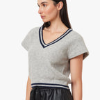 A woman modeling a light gray knit V-neck t-shirt with navy blue trim, paired with black leather pants, against a plain white background.