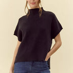 Woman wearing black ribbed mock neck short sleeve sweater paired with blue jeans, shown from chest up against cream backgrou…