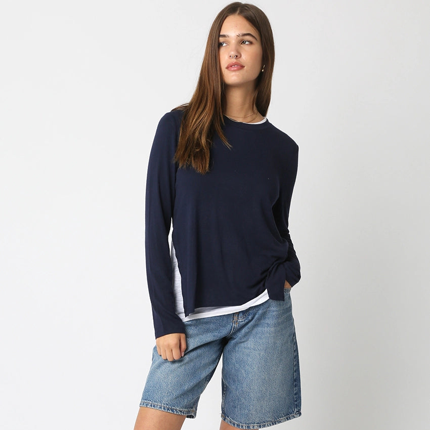 Woman wearing navy long-sleeve layered top with white trim over denim shorts, standing against white background