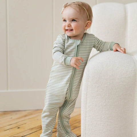 Toddler wearing sage green and cream striped zip-up sleeper, smiling while standing by white furniture