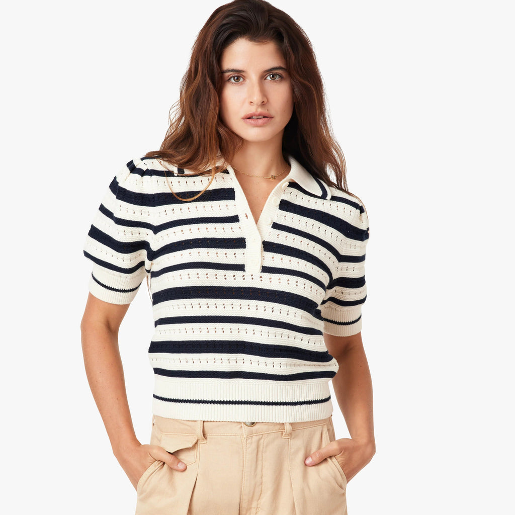 Woman wearing navy and cream striped polo sweater with short sleeves and beige trousers, standing against white background