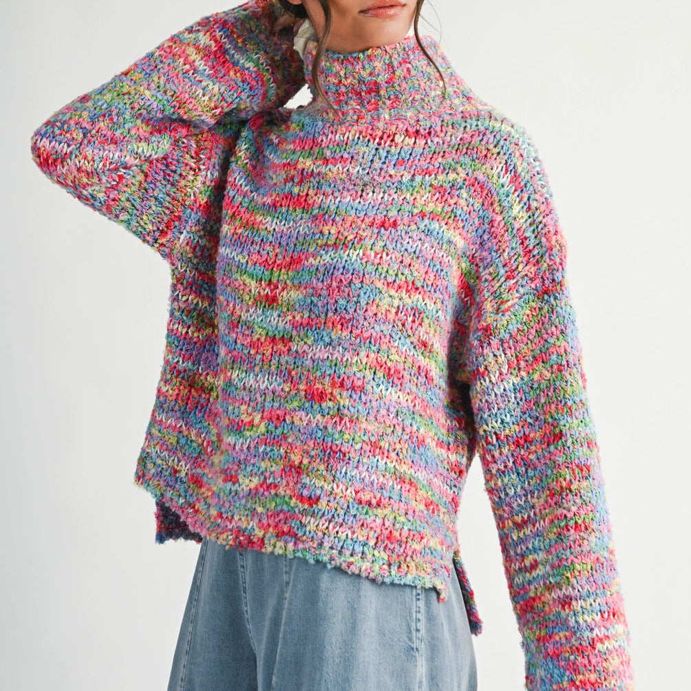 A woman wearing a chunky, multicolor knit turtle-neck sweater with long sleeves and light blue denim jeans, photographed against a plain light gray studio background.