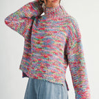 A woman wearing a chunky, multicolor knit turtle-neck sweater with long sleeves and light blue denim jeans, photographed against a plain light gray studio background.