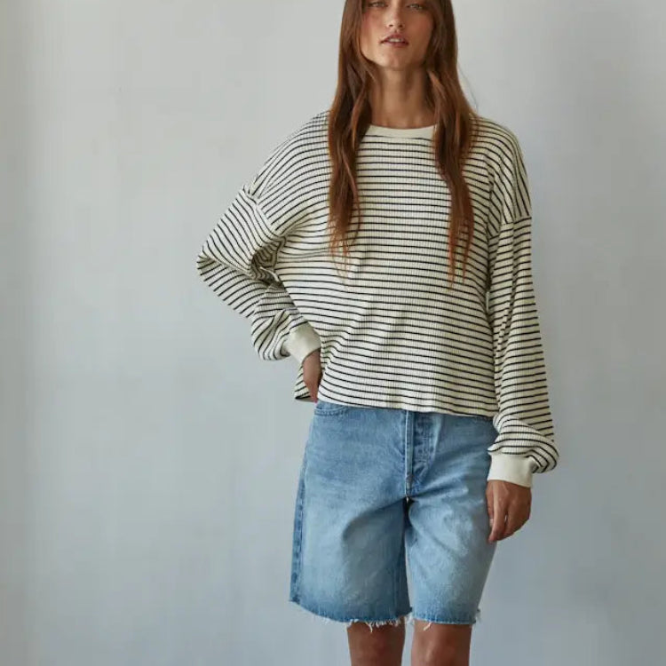 Woman wearing cream and navy striped long-sleeve cotton crew neck sweatshirt with blue denim shorts, standing against white …