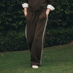 A woman stands outdoors on grass in front of a dense green hedge, wearing a dark brown sweatshirt and matching track pants with white side stripes, layered over a white collared shirt.