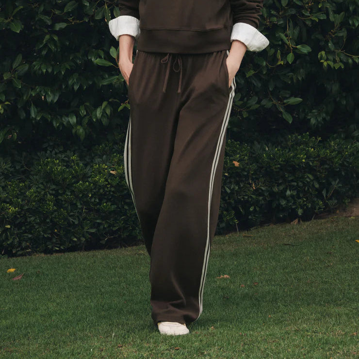 A woman stands outdoors on grass in front of a dense green hedge, wearing a dark brown sweatshirt and matching track pants with white side stripes, layered over a white collared shirt.