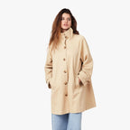 Woman wearing cream high neck trench coat with brown buttons over light blue jeans, standing against white background