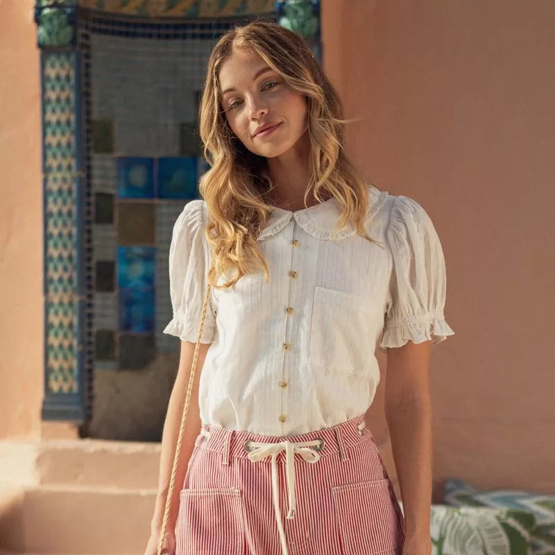 Woman wearing white puff sleeve blouse with pearl buttons and red striped pants, standing in front of ornate teal door